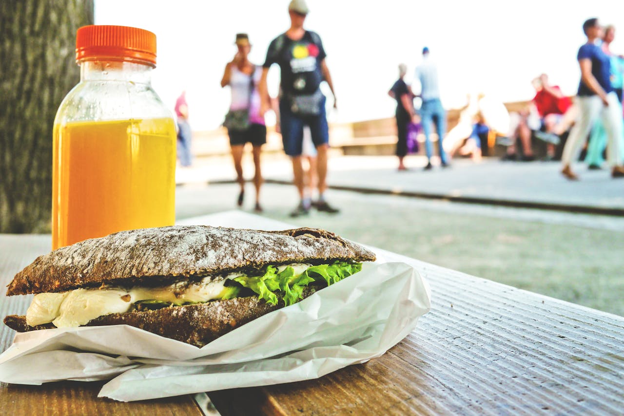 Close-up of a sandwich and orange juice on a table with people in the park.